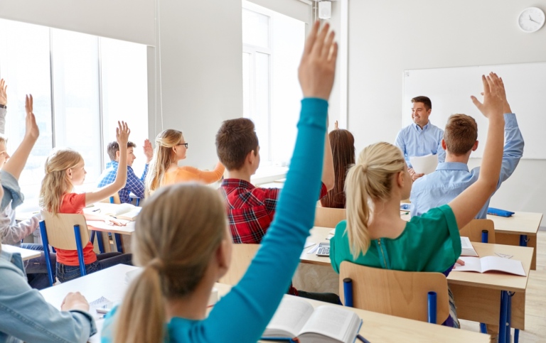  Students in a classroom all raising their hands, a male teacher smiling 