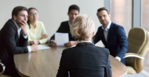 A woman in dark costume sits at a table with her back againt us, her collegues are looking at her