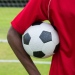  En mörkhyad fotbollsspelare håller i en fotboll på en fotbollsplan 
